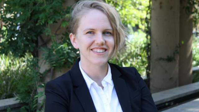 Smiling person with short blonde hair in a black blazer and white shirt, outdoor headshot