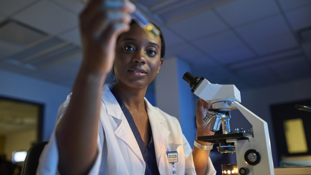 A UCLA Health researcher standing next to a microscope, holding a test tube