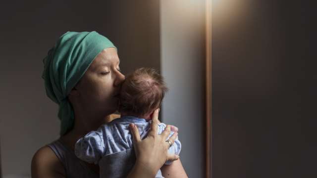 Woman in headscarf, fighting breast cancer while holding her newborn baby relaxing in cancer treatment hospital, patient standing next to hospital window. 