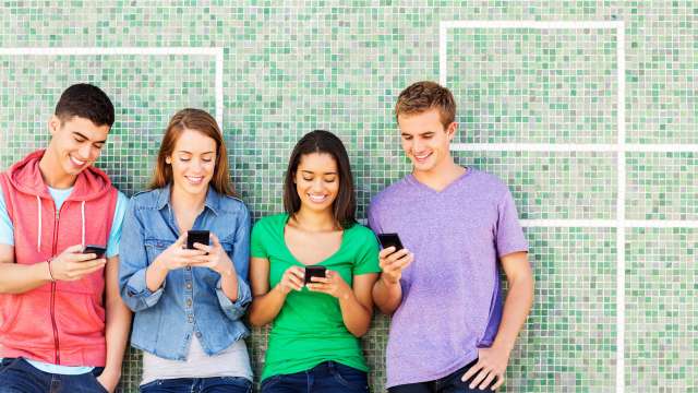 Teens against a wall looking at phones: four teens standing and using phones