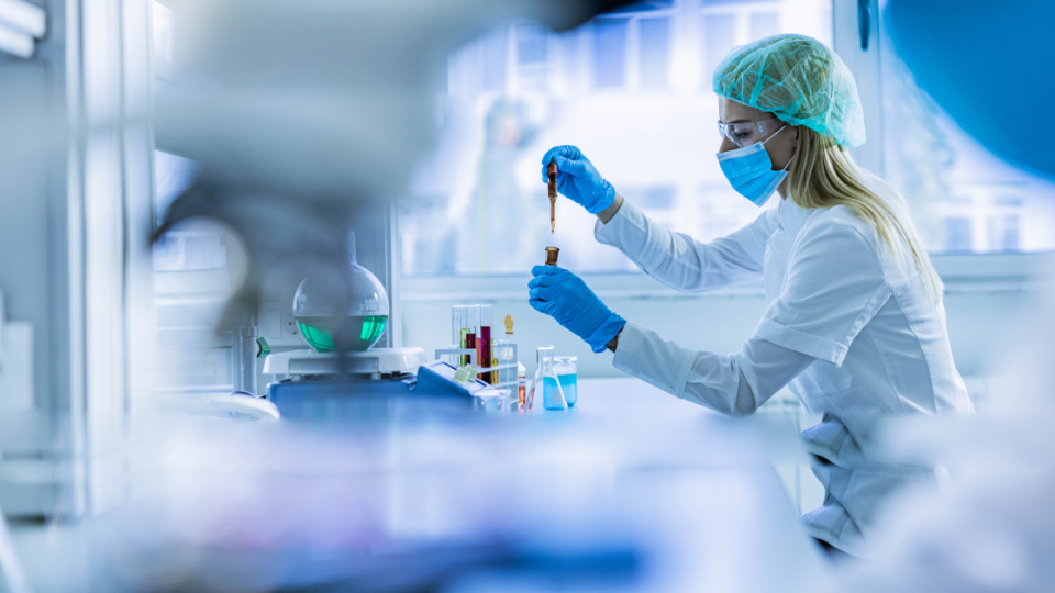 Woman wearing mask in a lab, holding testing materials such as beakers and chemicals.