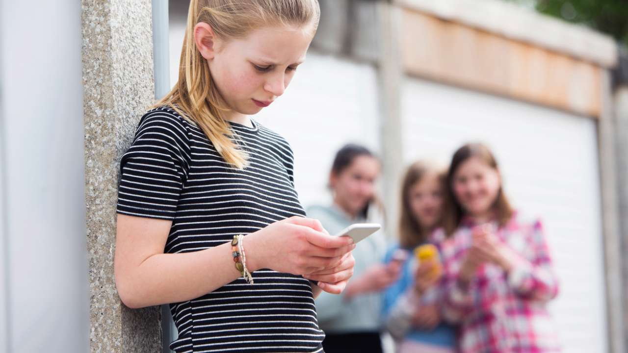 Sad girl looks down at her phone as a group of girls laughs at her in the background.