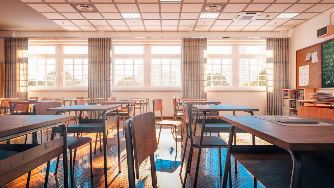Sunlit empty classroom with rows of desks and chairs, large windows with curtains, and a chalkboard on the wall.