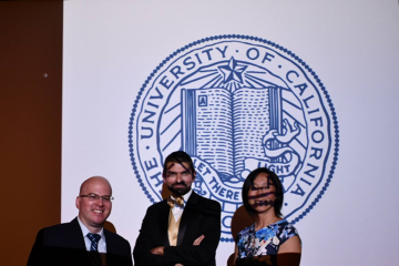 Three people in formal attire stand in front of a large University of California seal.