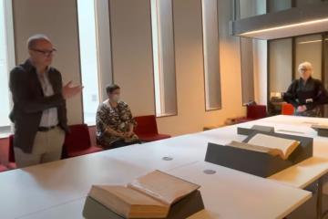 Three people in a room with large books displayed on angled stands atop a long white table. 