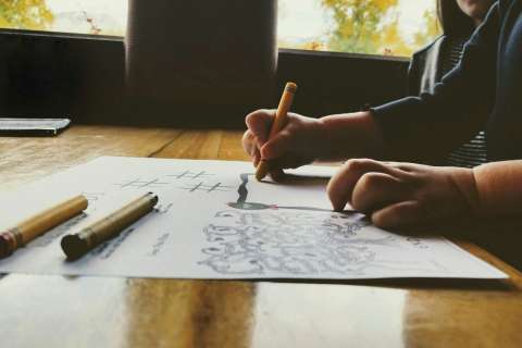 Hands sketching a drawing on paper with markers at a wooden table.