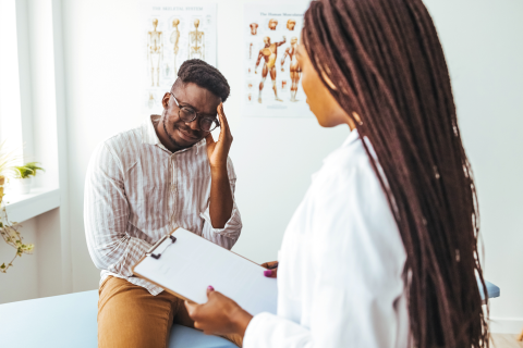 Patient struggling with migraine during a doctors visit. The doctor has a notepad and is writing down his symptoms for treatment.