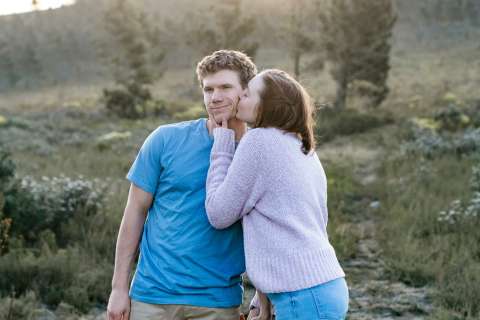 A young woman kisses her boyfriend on the cheek while they stand outdoors.