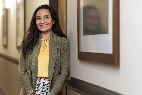 A person stands in a hallway, wearing a yellow shirt and green cardigan.