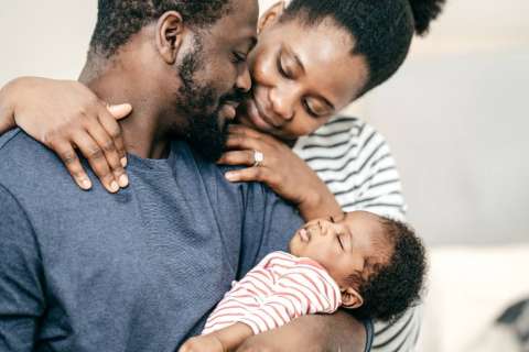 A family cuddles together, with a father, mother, and a sleeping baby.