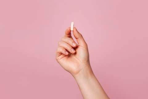 Hand holding small white pill against pink background.