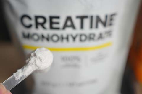 A scoop of creatine power is held in front of a bag of the supplement.
