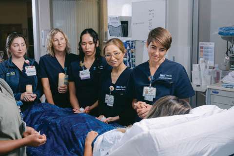 ICU nurses in scrubs support a patient in a hospital room.