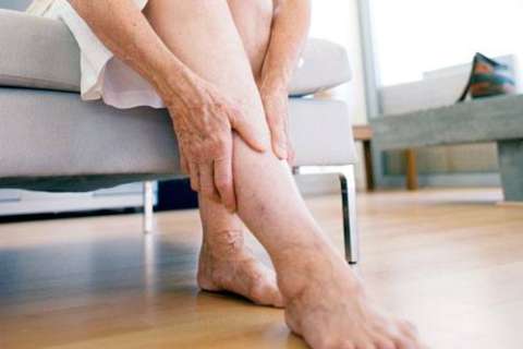 Person sitting on a chair, holding their ankle with one hand, bare feet resting on a wooden floor.