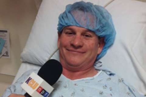 Lance Orozco in hospital gown and surgical cap lying in bed speaking to a microphone
