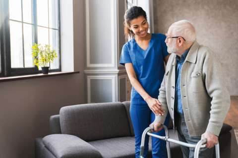 Portrait of an african young nurse helping old elderly disable man grandfather to walk using walker equipment in the bedroom. Senior patient of nursing home moving with walking frame and nurse support