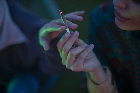 Person holding a cannabis cigarette, with another hand reaching towards it in dim lighting.