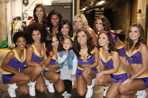  A group of ten cheerleaders in purple and gold uniforms crouches around a small child, posing for a picture in an indoor setting.