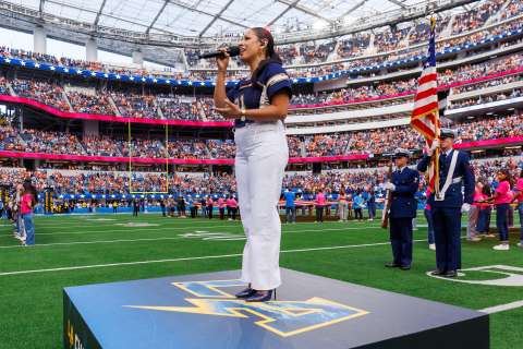 A woman with a microphone stands on the podium on the field in front of a larger crowd before a Los Angeles Chargers game.