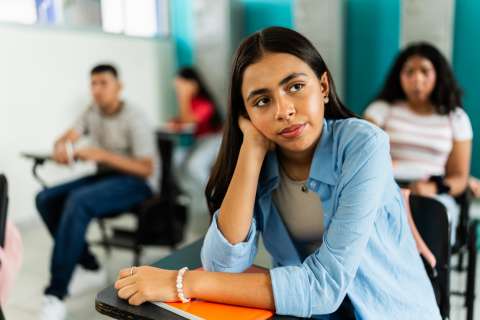 Student resting chin on hand, holding notebook, looking thoughtful in classroom.