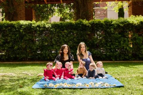 Two new moms and their identical triplets sit on a blanket together.