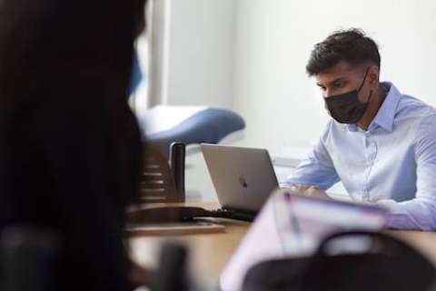 Vikram Krishna, wearing a black face mask and a light blue shirt, works on a laptop at a wooden desk.
