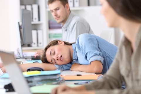 Woman sleeping with head on desk in classroom while coworkers look at her with concerned expressions.