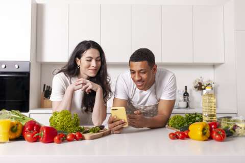 A smiling couple looks at a smartphone while unpacking healthy groceries in their kitchen.
