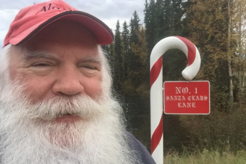 Man with white beard at No. 1 Santa Claus Lane sign shaped like a candy cane, standing in front of trees outdoors.