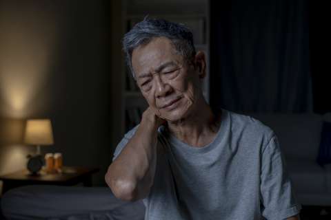Older man holding his neck with a pained expression in a dim bedroom.