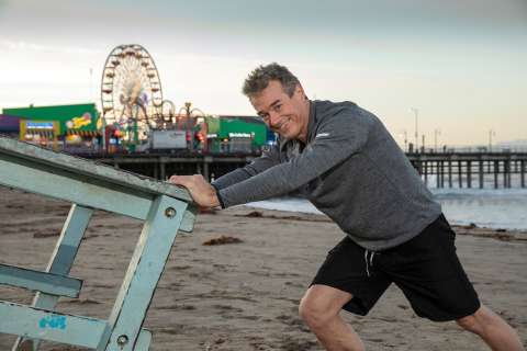 Man pushing a lifeguard stand on the beach with a pier and ferris wheel in the background.