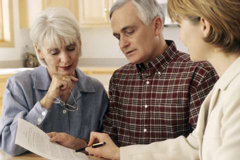 A senior couple in their home talking with an agent, all referencing a printed paper or form.