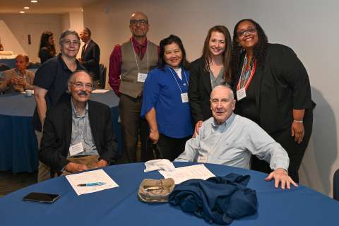 Group of seven people posing together at a conference, smiling around a table.