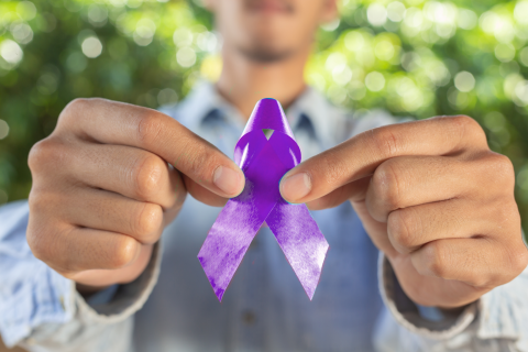 Close-up of a man's hands gently holding a purple ribbon, symbolizing prostate cancer awareness, against a soft, neutral background. The image conveys support, care, and advocacy for prostate cancer education and research.