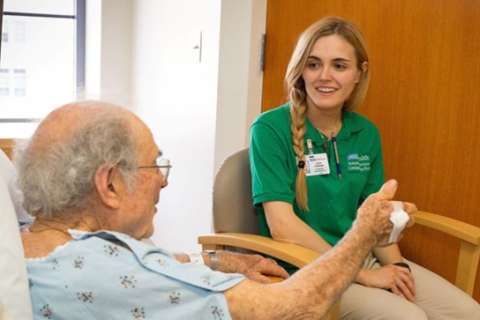 A young woman speaking with an older patient resting in his bed