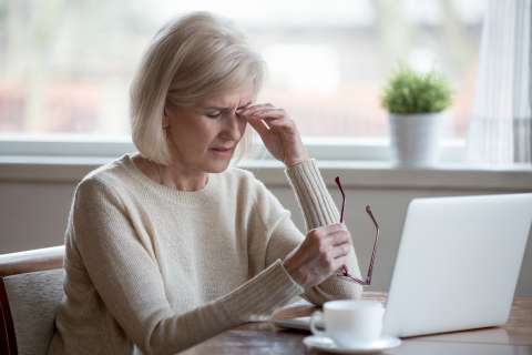 A woman sits in front of a laptop with her eyes closed, touching her eyelid.