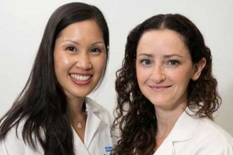 A portrait of two female medical professionals, one with straight hair and one with curly hair, both smiling and wearing white lab coats.