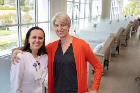 Lori Jordan, in an orange cardigan, and a doctor, in a white coat, stand arm-in-arm and smile in a brightly lit clinic hallway.