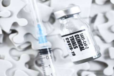  A close-up of a syringe and a glass vial labeled "SARS-CoV-2 CLINICAL TRIALS COVID-19" resting on a background of white puzzle pieces.