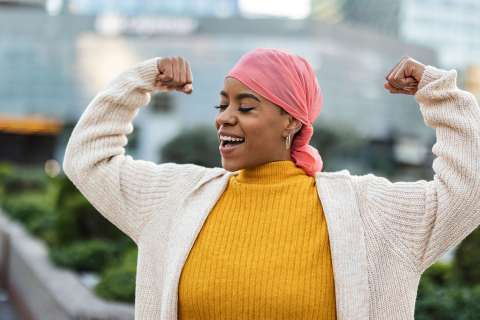 Person outdoors smiling and flexing both arms, wearing a pink headscarf and yellow sweater