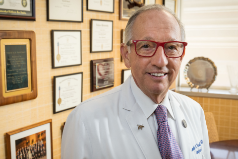 Ronald W. Busuttil, MD, PhD standing in front of a yellow wall with many picture frames, smiling and looking at the camera
