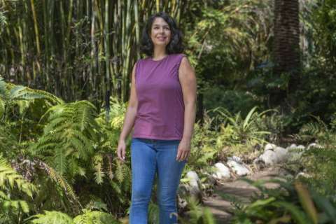 Caitlin Musick in a purple shirt and blue pants stands in a forest, looking directly at the viewer with a slight smile.