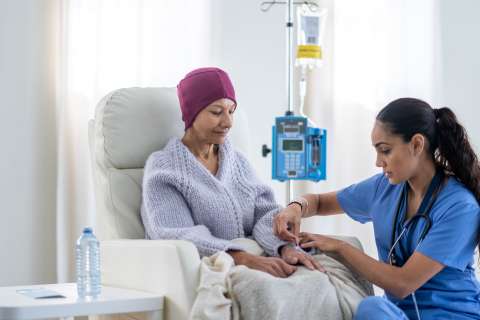 Woman in headscarf seated receiving IV infusion while nurse checks her hand