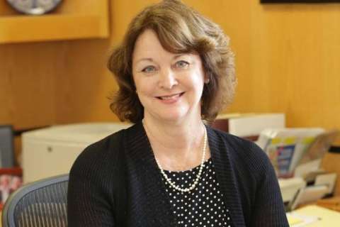 Judy Gasson seated at a desk, wearing a black cardigan and pearl necklace.
