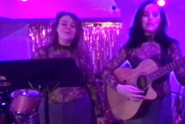  Two women perform under purple stage lights, one singing at a mic stand, the other playing an acoustic guitar.