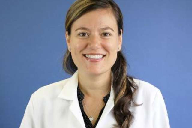 Person wearing a white lab coat over a dark shirt, posed against a solid blue background.