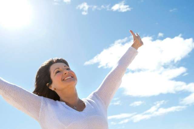 A smiling woman with dark hair, wearing a white sweater, stands outdoors with her arms raised joyfully toward the bright blue sky and sun.