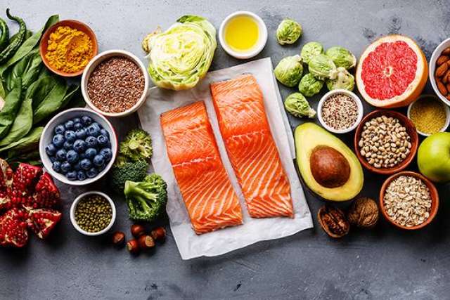 Assorted healthy foods including salmon fillets, avocado, grapefruit, nuts, seeds, berries, and leafy greens arranged on a gray surface.
