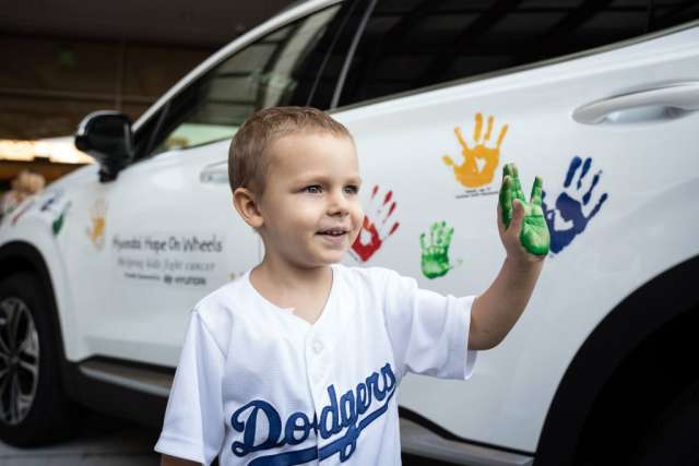A young boy in a "Dodgers" shirt, smiling, shows his green-painted hand while standing next to a white car covered in colorful handprints.