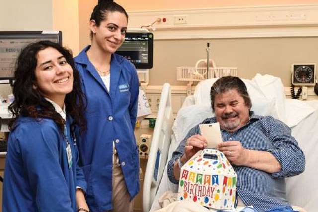 Hospital patient sitting in a bed holding a card, with a colorful gift bag that says ‘Happy Birthday’ on the table, and two people in blue uniforms standing nearby.